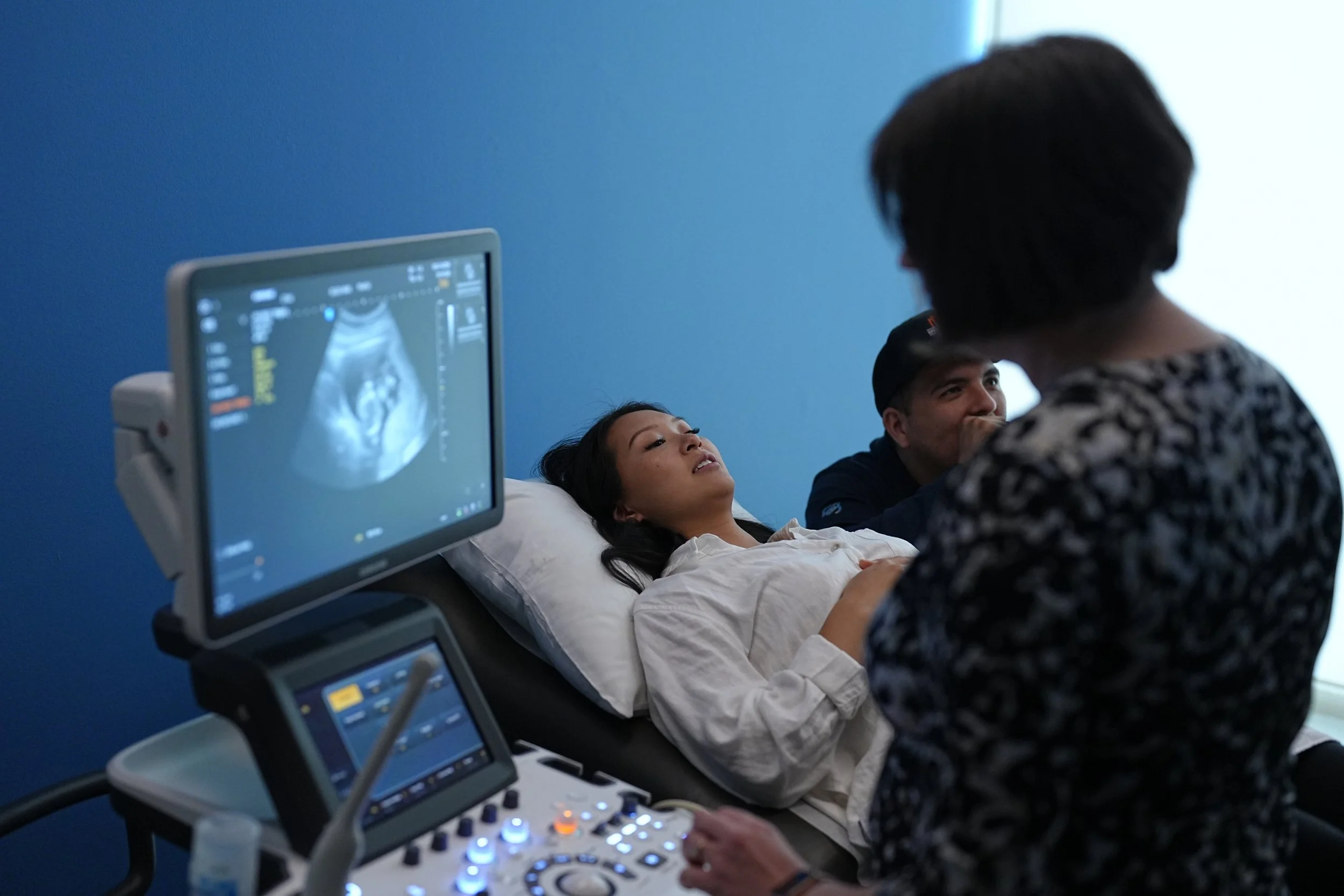 Medical professional performing an ultrasound on a pregnant woman lying in a hospital bed, with a monitor displaying the ultrasound image and a partner sitting beside them.
