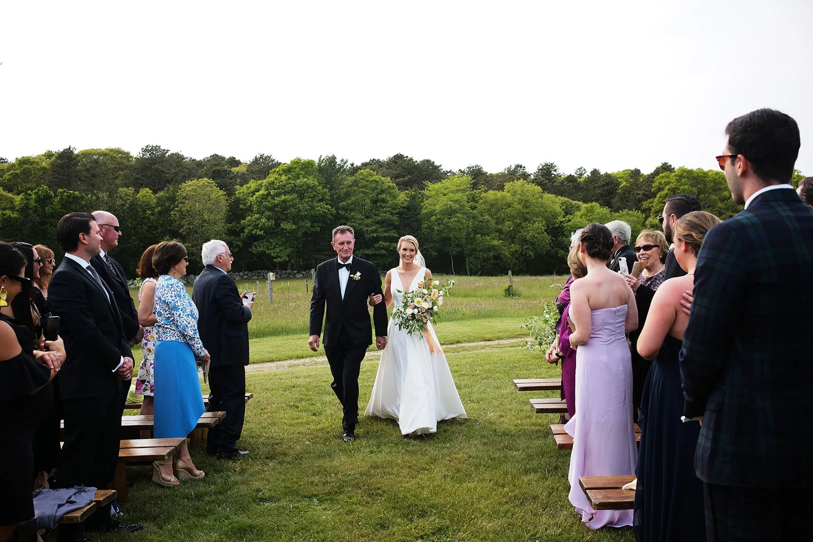 Bride walking down the aisle with her father as guests watch at an outdoor wedding ceremony with green trees in the background.