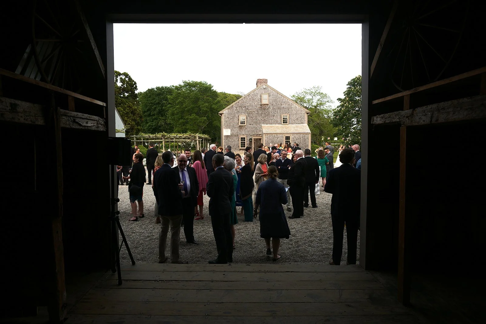 People gathered outdoors at a social event, seen from inside a barn toward a rustic wooden house and trees.