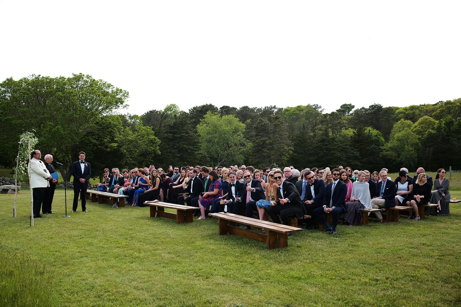 Outdoor wedding ceremony with guests seated on wooden benches on a grassy field surrounded by trees, officiant and groom at the front.