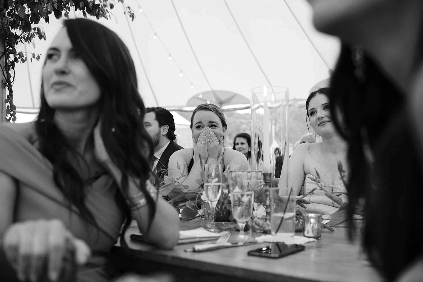 A group of women and men seated at a table outdoors under a tent, with some women appearing emotional or touched, as they participate in a celebration or event.