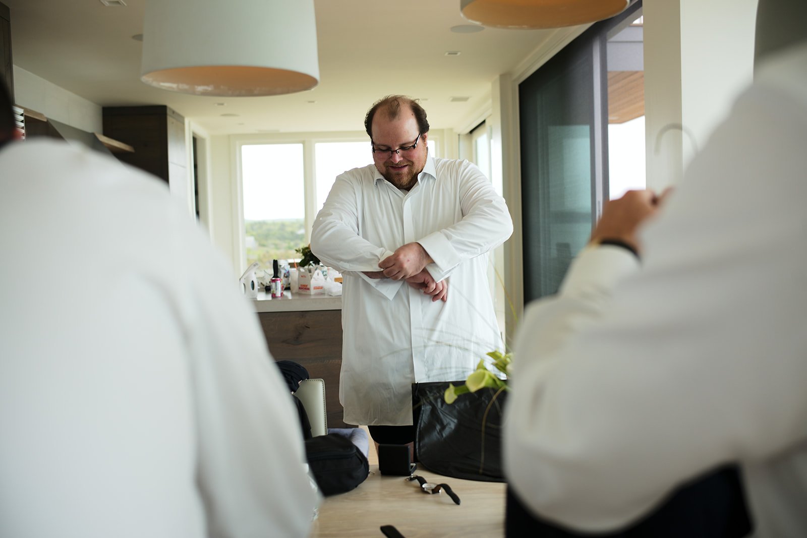 Man in white shirt adjusting his sleeves in a bright room with large windows, seen from behind by two other people also in white shirts.