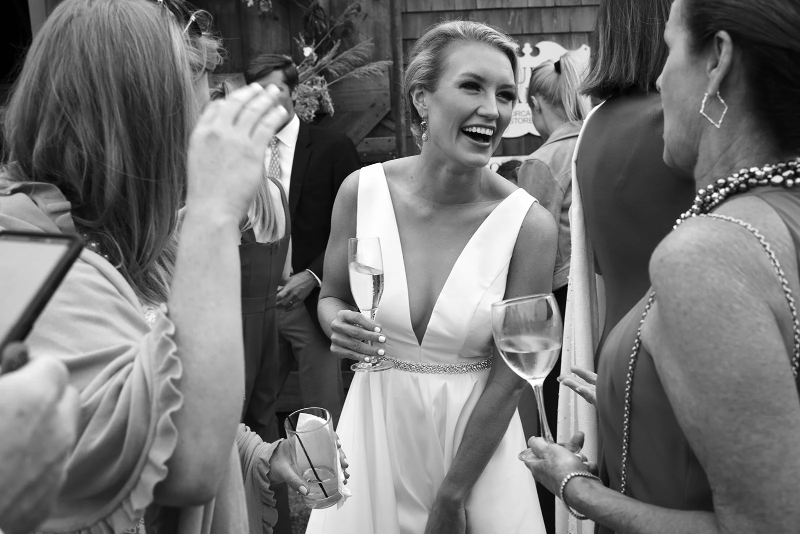 A woman in a white dress holding a glass of champagne, smiling and talking with a group of women at a social gathering.