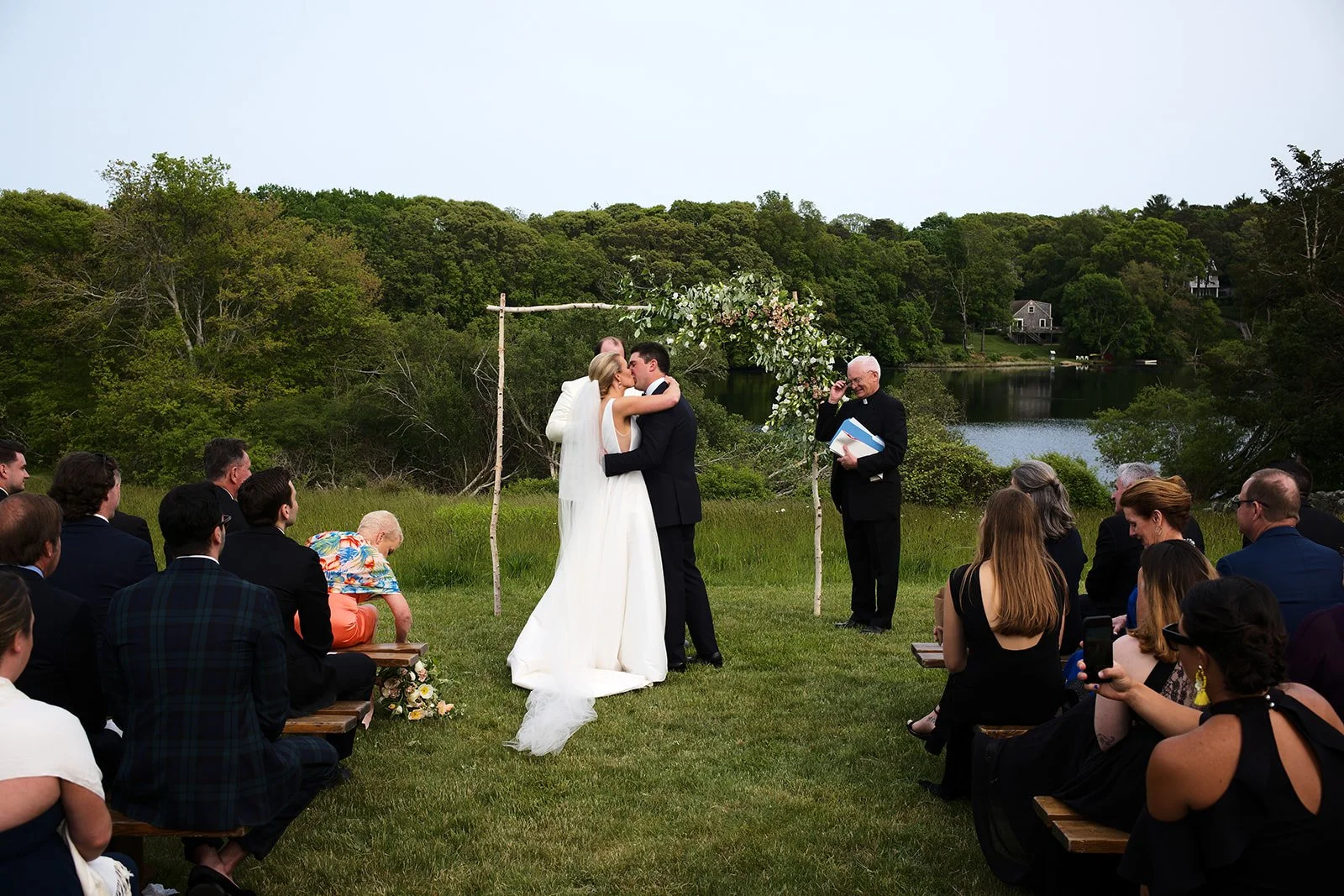 A wedding ceremony outdoors by a lake with a green landscape, where a couple is kissing under an arch decorated with flowers, surrounded by seated guests.