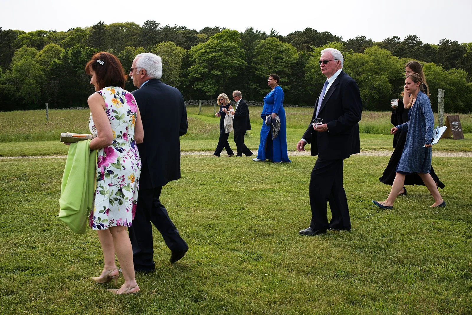 People walking on a grassy field during an outdoor event, with a wooded area in the background.
