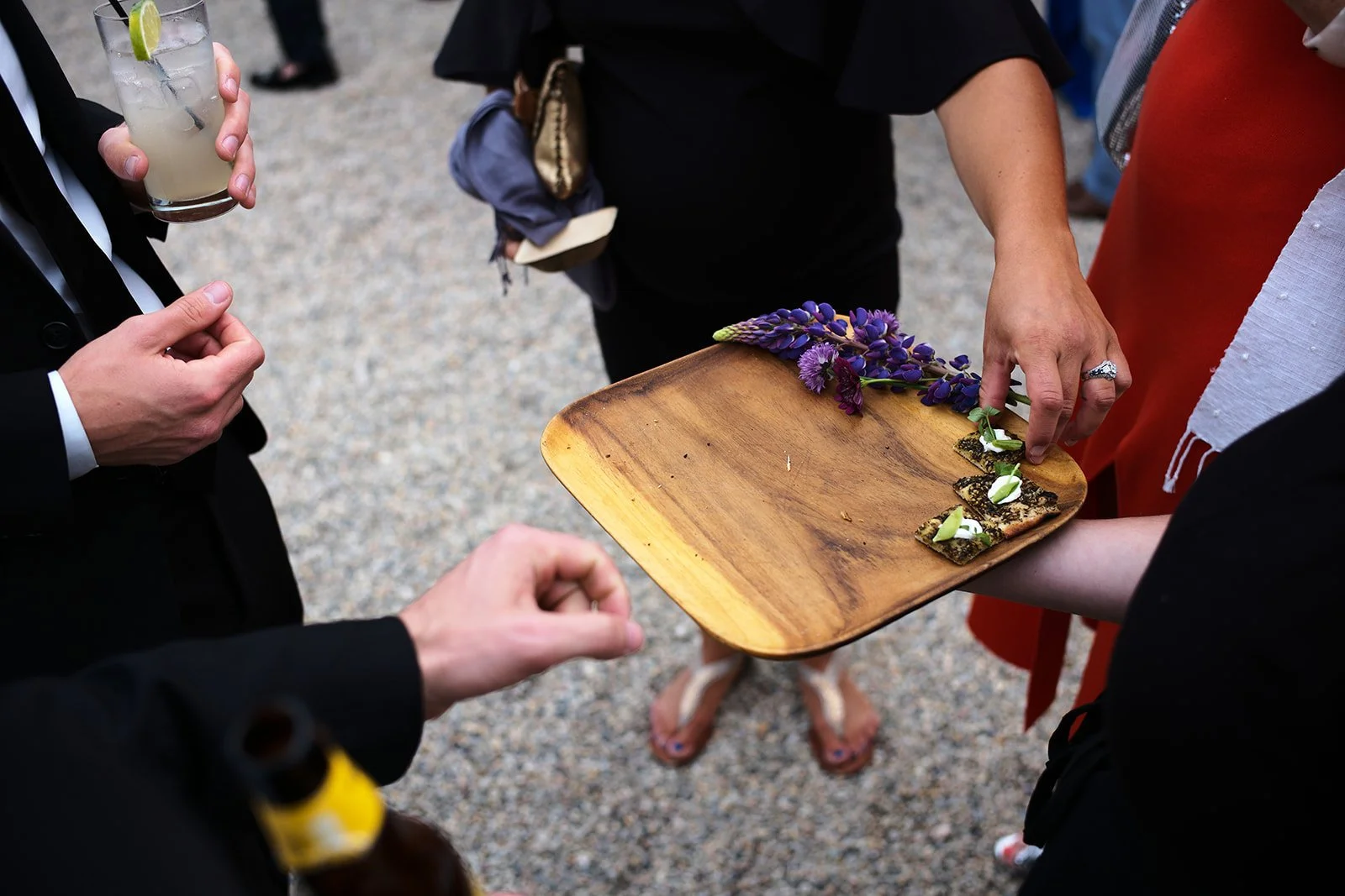 People at a social gathering are being served food on a wooden tray. The tray has purple flowers and small pieces of sushi or appetizers with green and white garnishes. One person is handing the tray to another person, while others are holding drinks