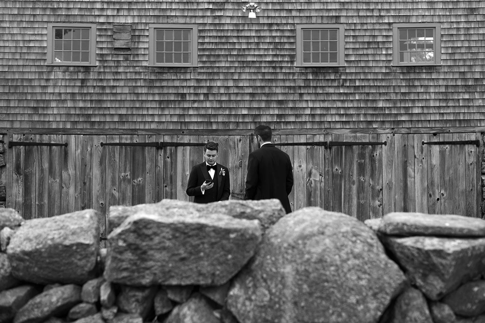 Two men in tuxedos standing in front of a wooden fence, one looking at his phone, behind a stone wall, with a building in the background.