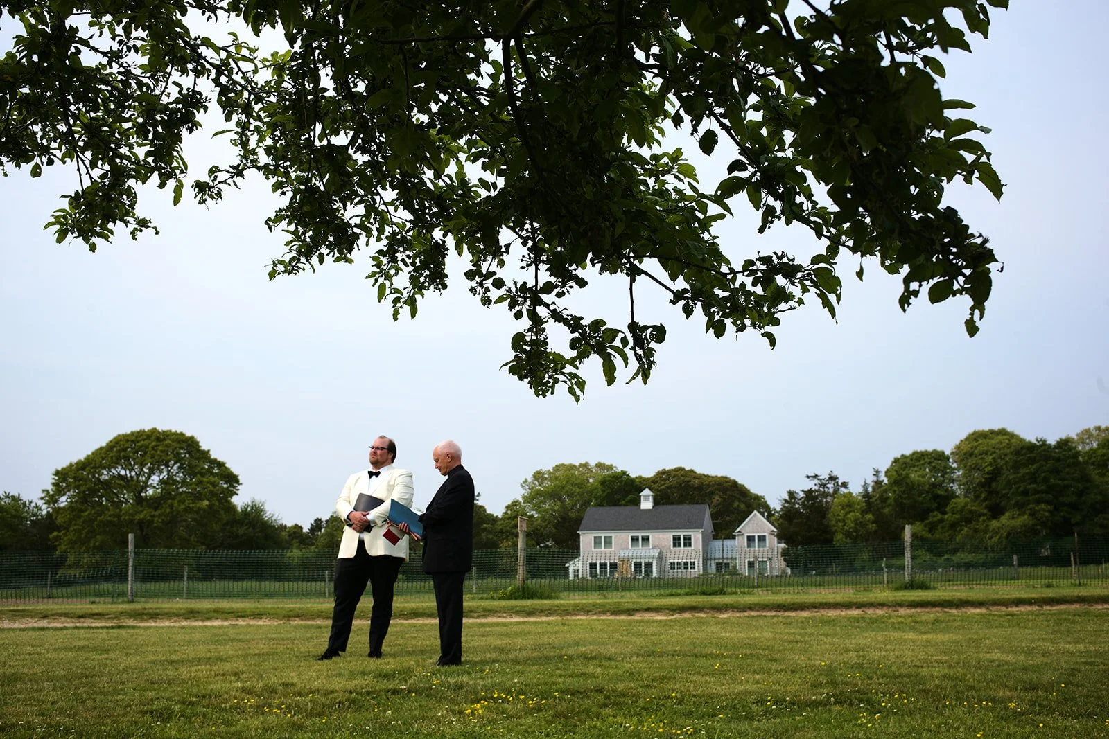 Two men standing on a grassy field, dressed in formal attire, holding books and papers, with a house and trees in the background under a clear sky.
