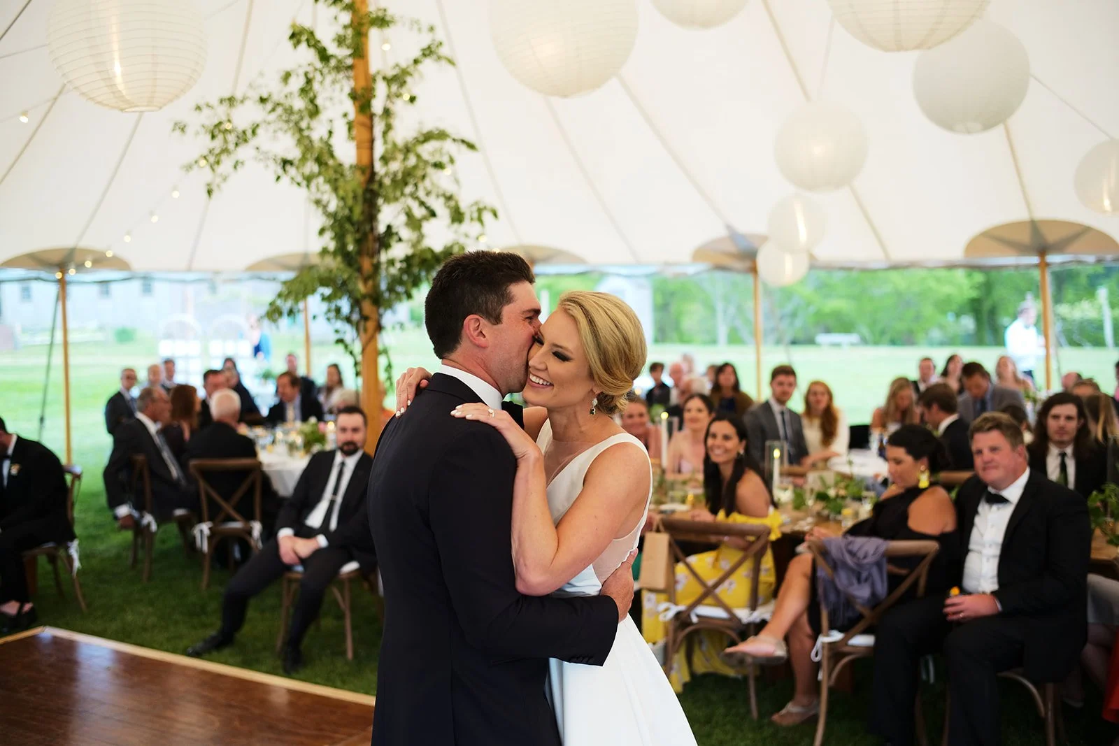 A bride and groom sharing a dance at their wedding reception inside a large tent, with seated guests watching in the background.