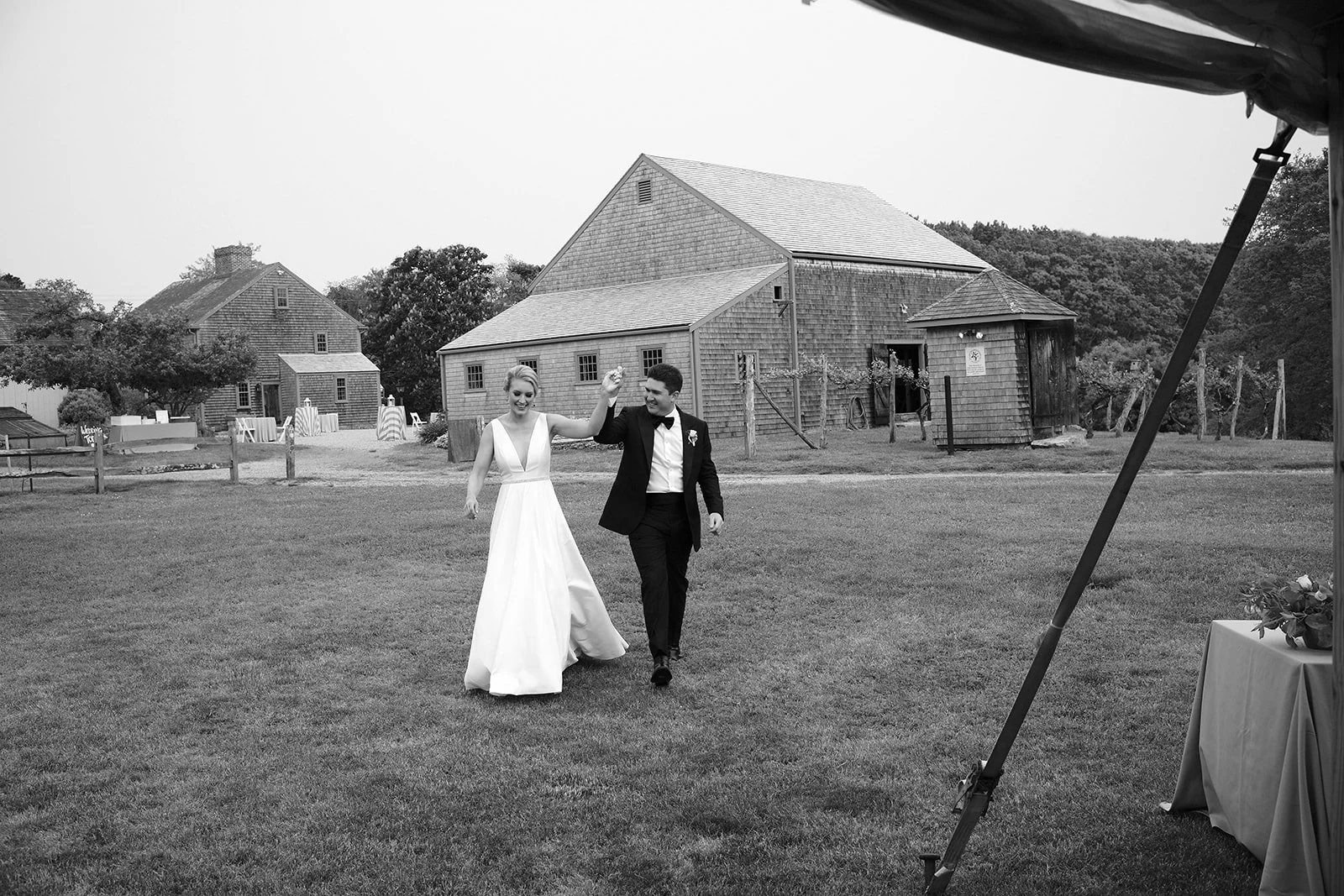 A newlywed couple walking on a grassy field, holding hands and smiling, with rustic wooden buildings in the background, during daytime.