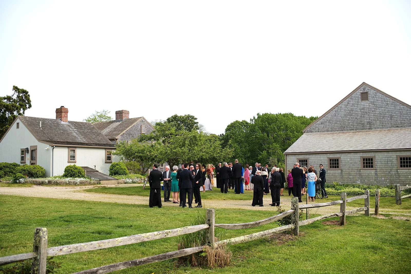 Group of people dressed in formal attire gathering outdoors on a grassy area near farmhouse-style buildings with trees in the background.