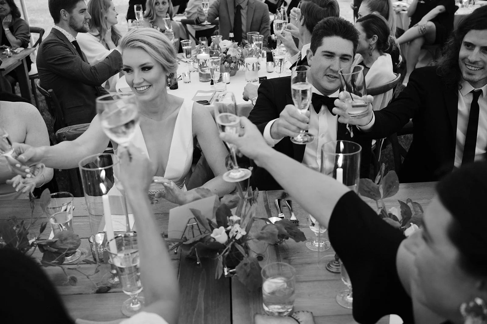Group of people celebrating at a wedding reception, raising champagne glasses in a toast, with a woman in a white dress and a man in a tuxedo at the center of the image.