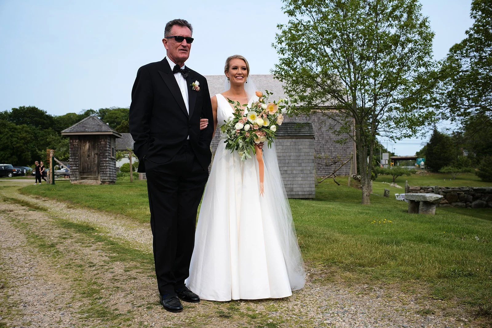 A bride in a white wedding dress holding a bouquet of flowers, standing next to a man in a black tuxedo with sunglasses, in an outdoor setting with trees and small buildings.
