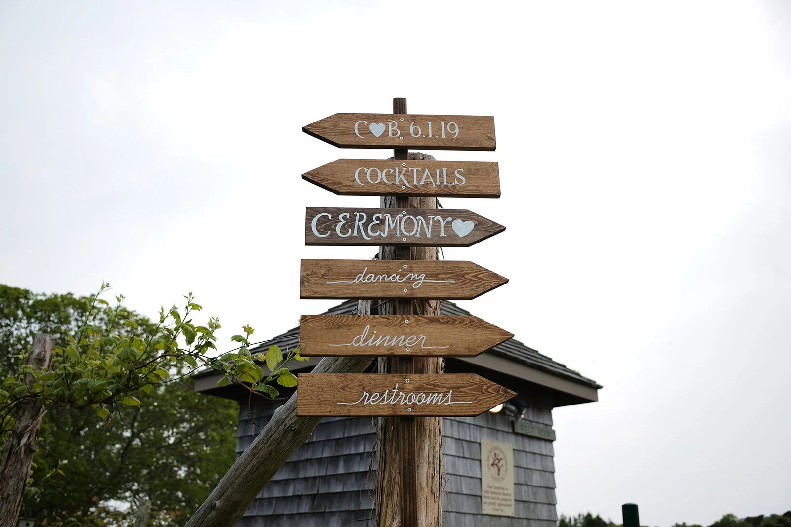 Wooden directional signpost with arrows pointing to various wedding event locations, including cocktails, ceremony, dancing, dinner, and restrooms, standing in front of a rustic building on a cloudy day.