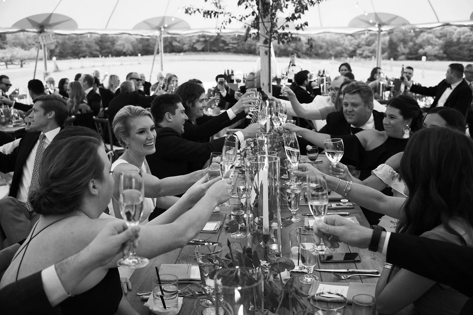 Black and white photograph of people in formal attire raising glasses in a toast at an outdoor wedding reception under a large tent.