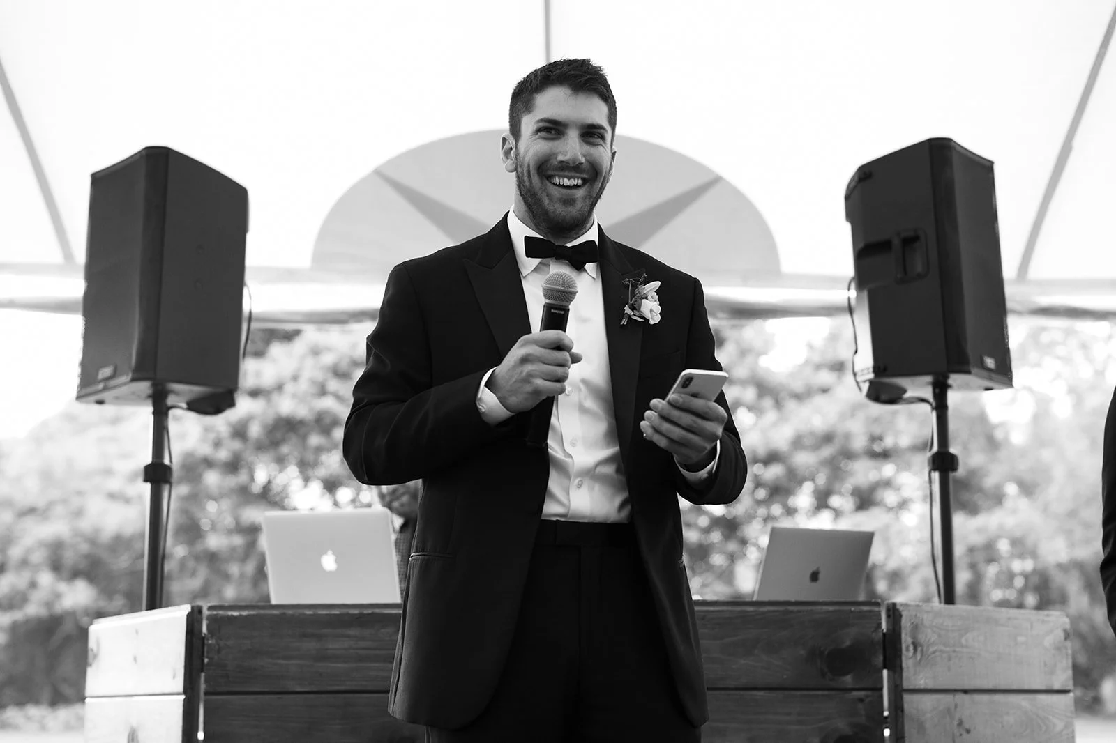 A man in a tuxedo holding a microphone and a smartphone, smiling at an outdoor event with speakers and laptops in the background.