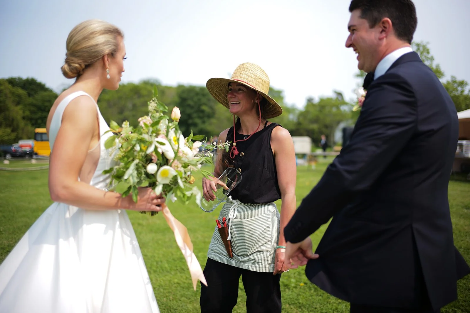 A woman in a white wedding dress holding a bouquet of flowers, a man in a tuxedo, and a woman wearing a straw hat and black outfit standing outdoors on a grassy field, smiling and talking.