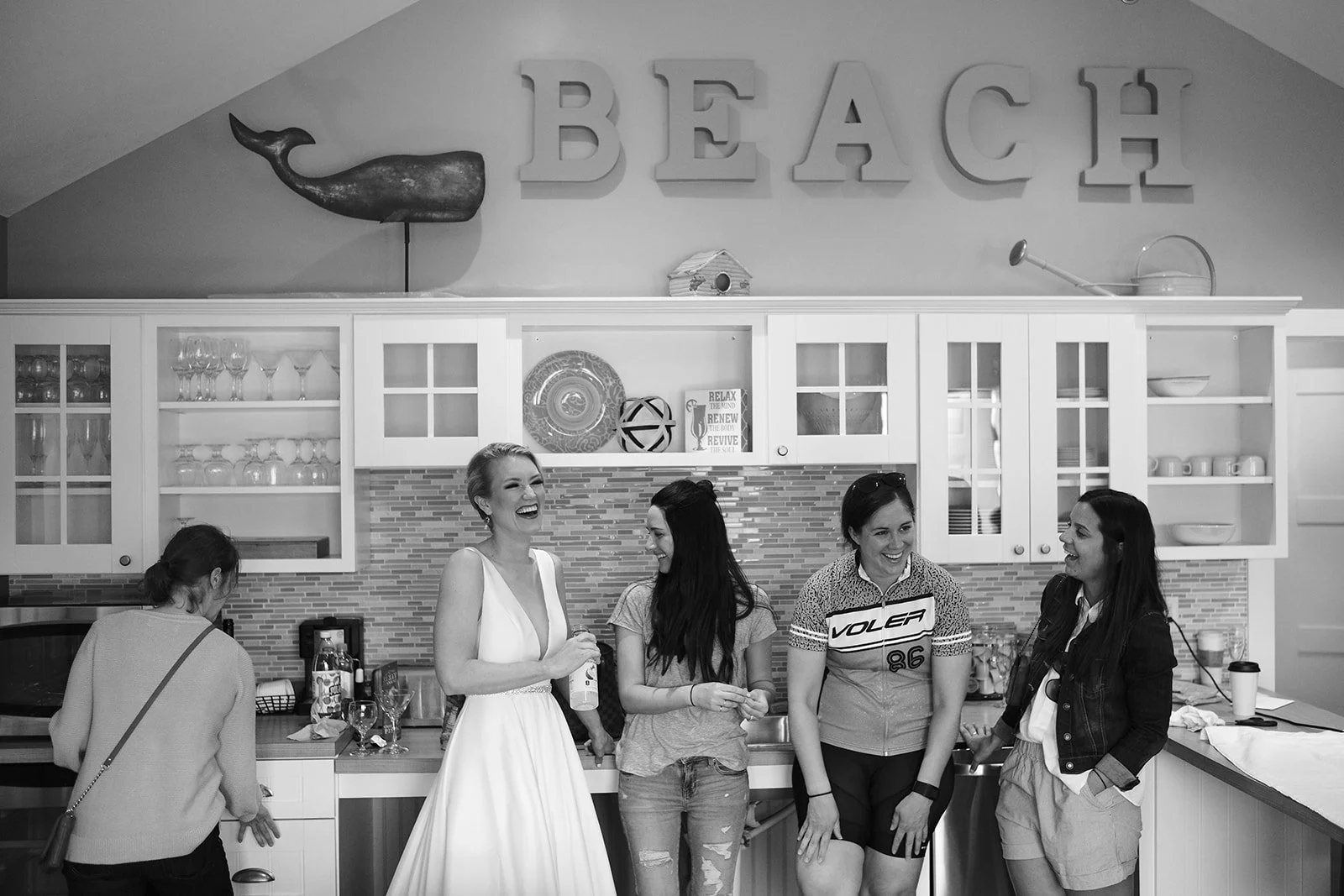 Group of five women standing and laughing in a kitchen with 'BEACH' sign and whale wall art above