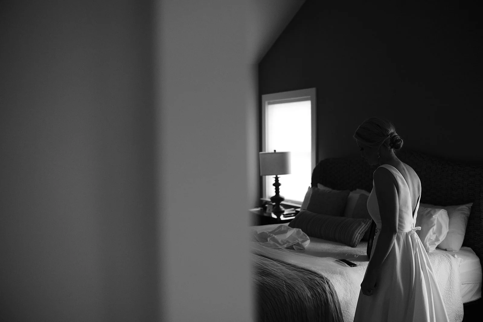 A woman in a white dress stands in a bedroom, looking down, with natural light coming through a window behind her. The room has pillows on the bed, and a nightstand with a lamp.