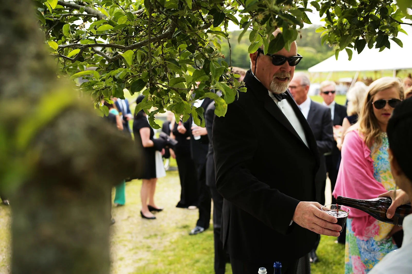 Man in tuxedo pouring a drink at an outdoor social event, surrounded by people in semi-formal attire and sunglasses, under a tent or canopy.