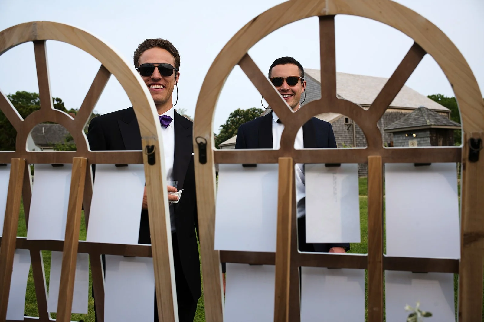 Two men in suits and sunglasses standing outside behind a wooden decorative screen, smiling, with houses and trees in the background.