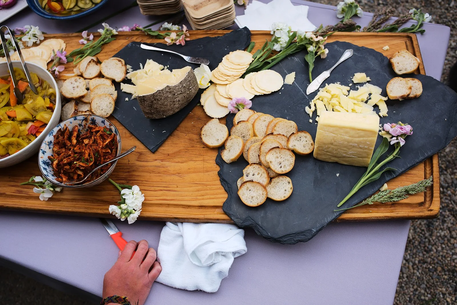 A cheese and bread platter set on a wooden board with flowers and a tablecloth beneath, featuring sliced bread, various cheeses, crackers, a bowl of pickled vegetables, and a small bowl of shredded meat.