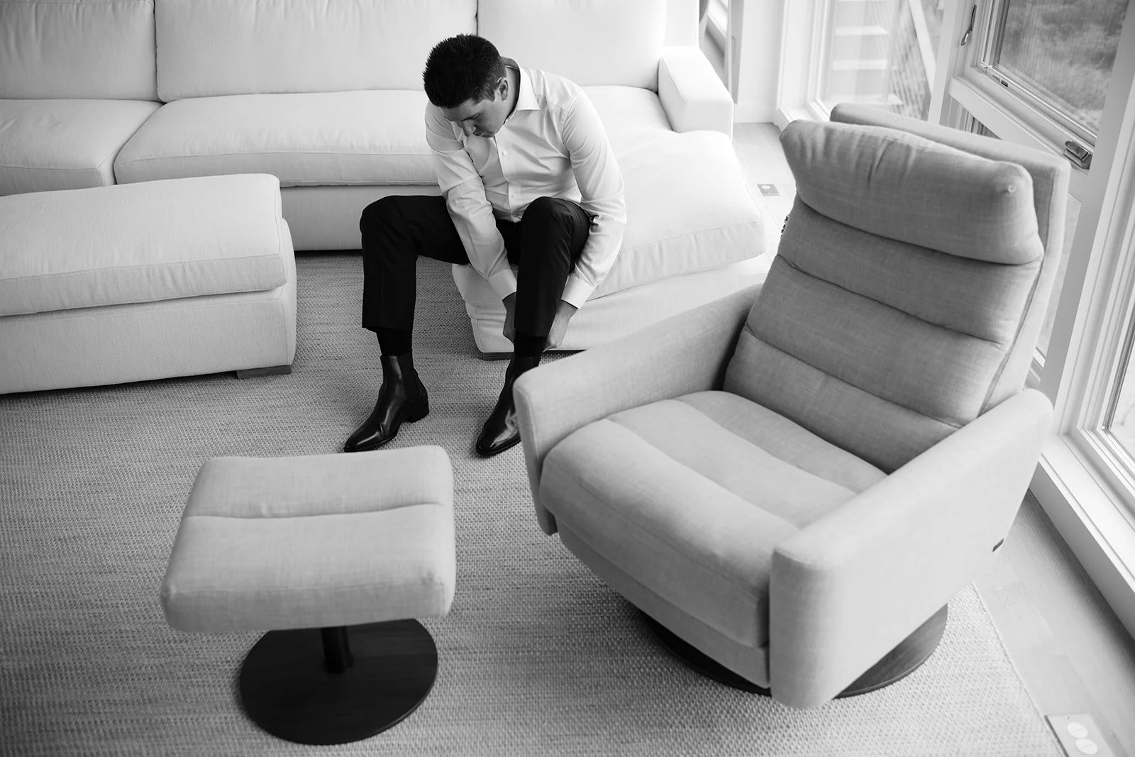 A man in formal attire is sitting on a light-colored sofa, adjusting his black shoes in a cozy, well-lit living room with large windows.