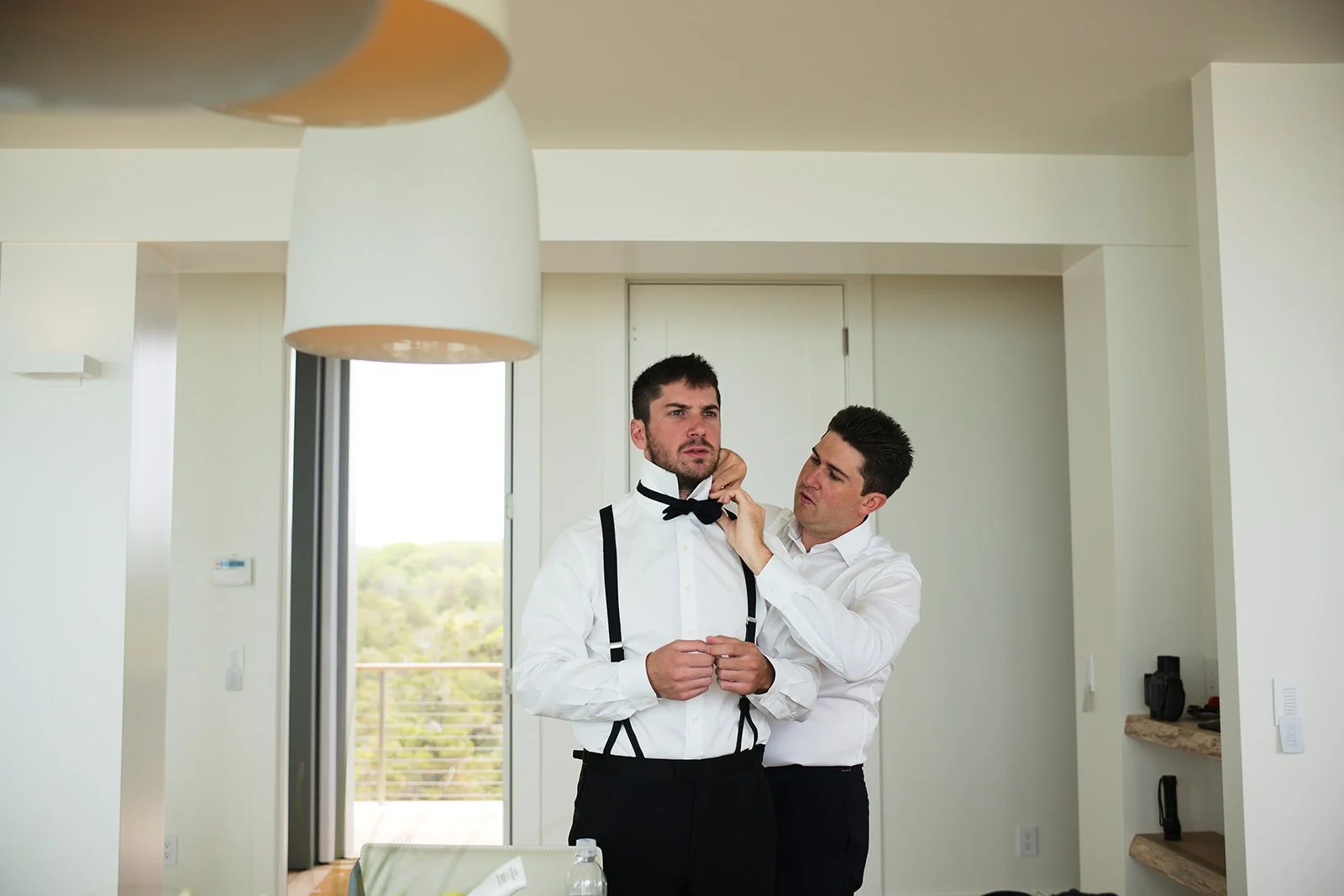 Man in a white shirt with suspenders standing as another man adjusts a bowtie in a modern room with large windows.