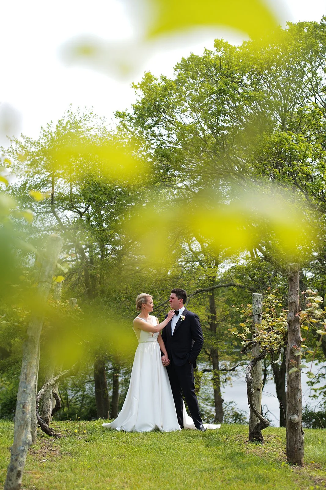 Couple in wedding attire, woman in white gown and man in black tuxedo, standing on grass under green trees, holding hands, outdoors during daytime.