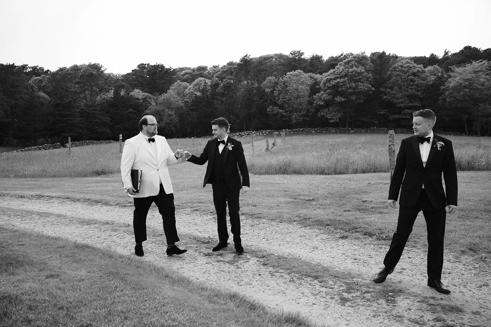 Black and white photo of three men in suits standing outdoors in a field with trees in the background. One man on the left is wearing a white blazer and holding a book, the middle man is handing a small object to him, and the third man on the right i
