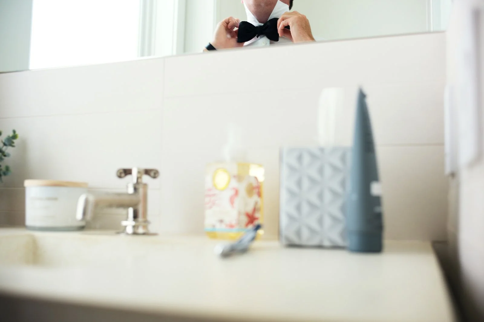 A man adjusting a black bow tie in front of a mirror bathroom sink countertop with toiletries and a green plant.