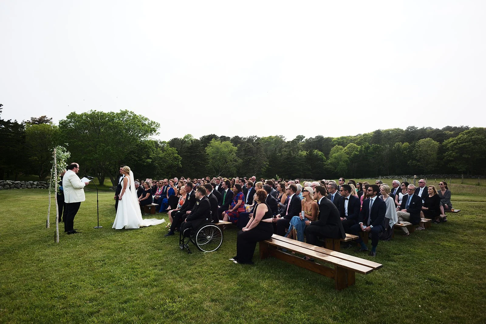 Outdoor wedding ceremony with bride and groom facing officiant, seated guests, and scenic green landscape.