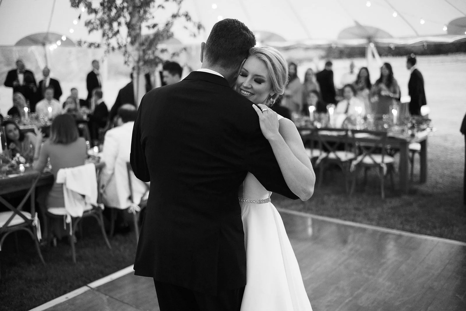 A bride and groom sharing a dance at their wedding reception, with guests seated at tables in the background.