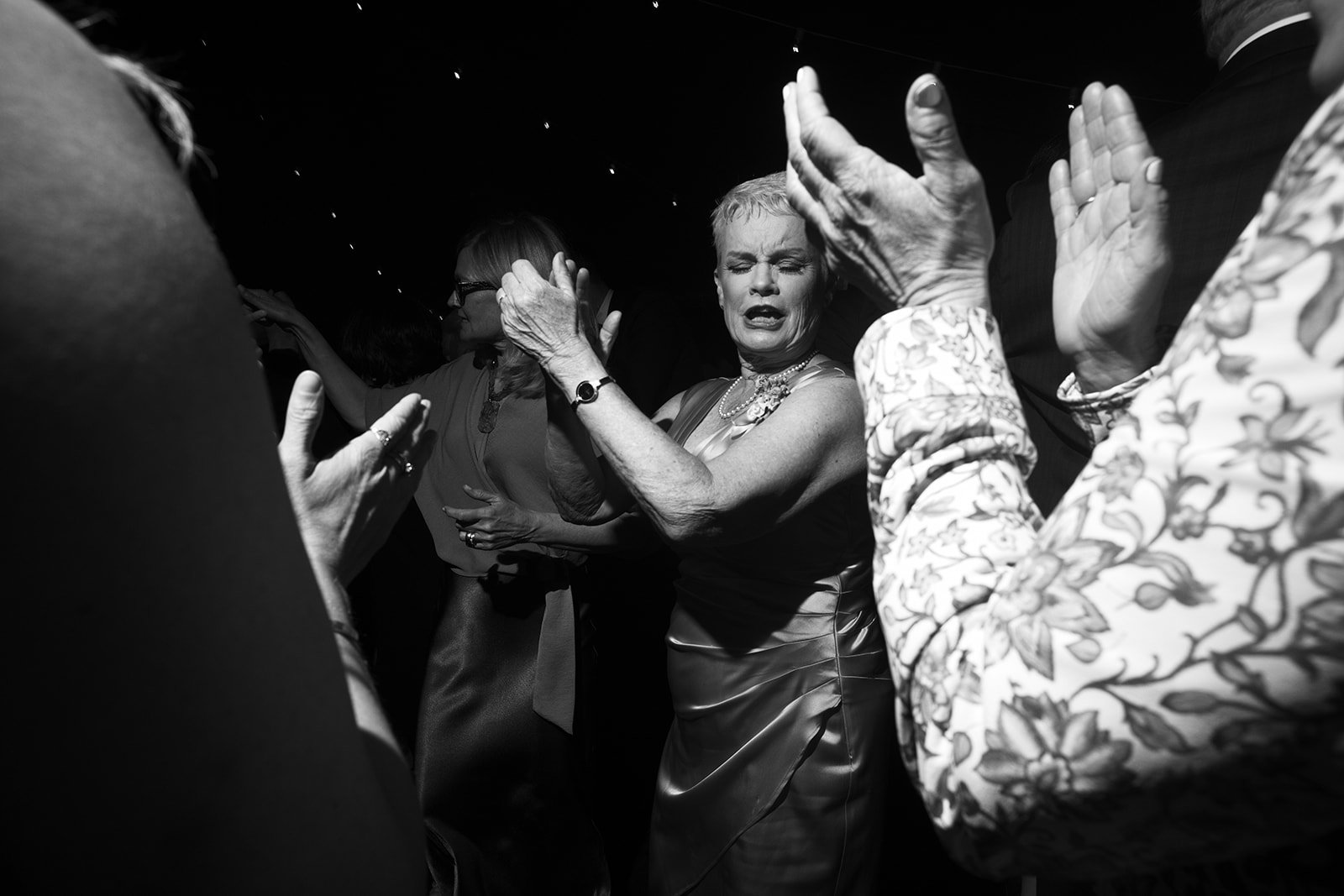 An elderly woman with short hair and a satin dress appears emotional with her eyes closed and hands raised, surrounded by other people clapping in a dark setting.