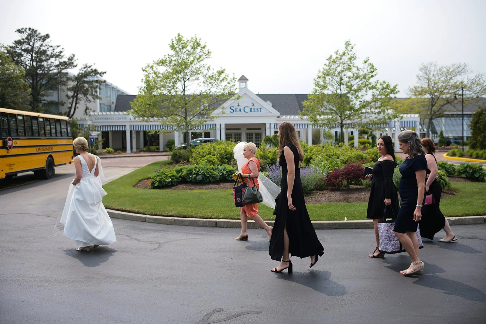 A bride in a white wedding gown walking on a paved road, followed by several women dressed in black, possibly bridesmaids, near the Sea Crest hotel with a yellow bus parked nearby.