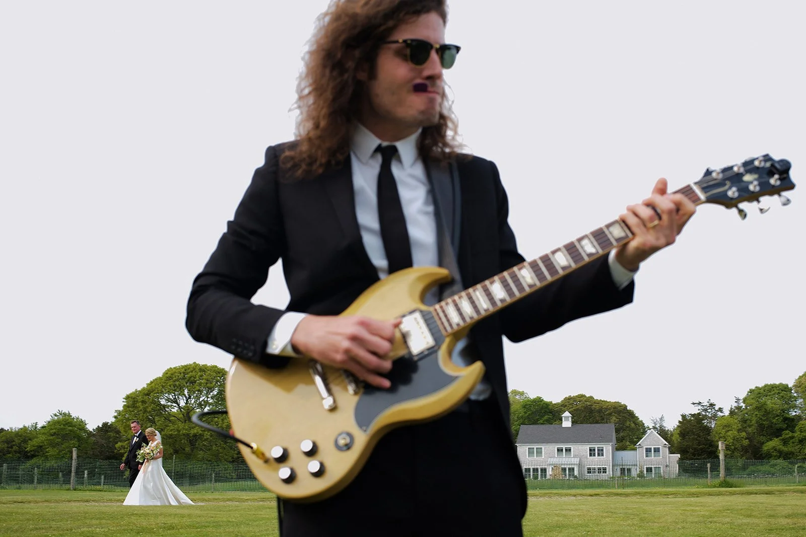 A man in a black suit, white shirt, black tie, and sunglasses playing an electric guitar outdoors. In the background, a bride and groom are walking on a grassy field near some houses and trees.