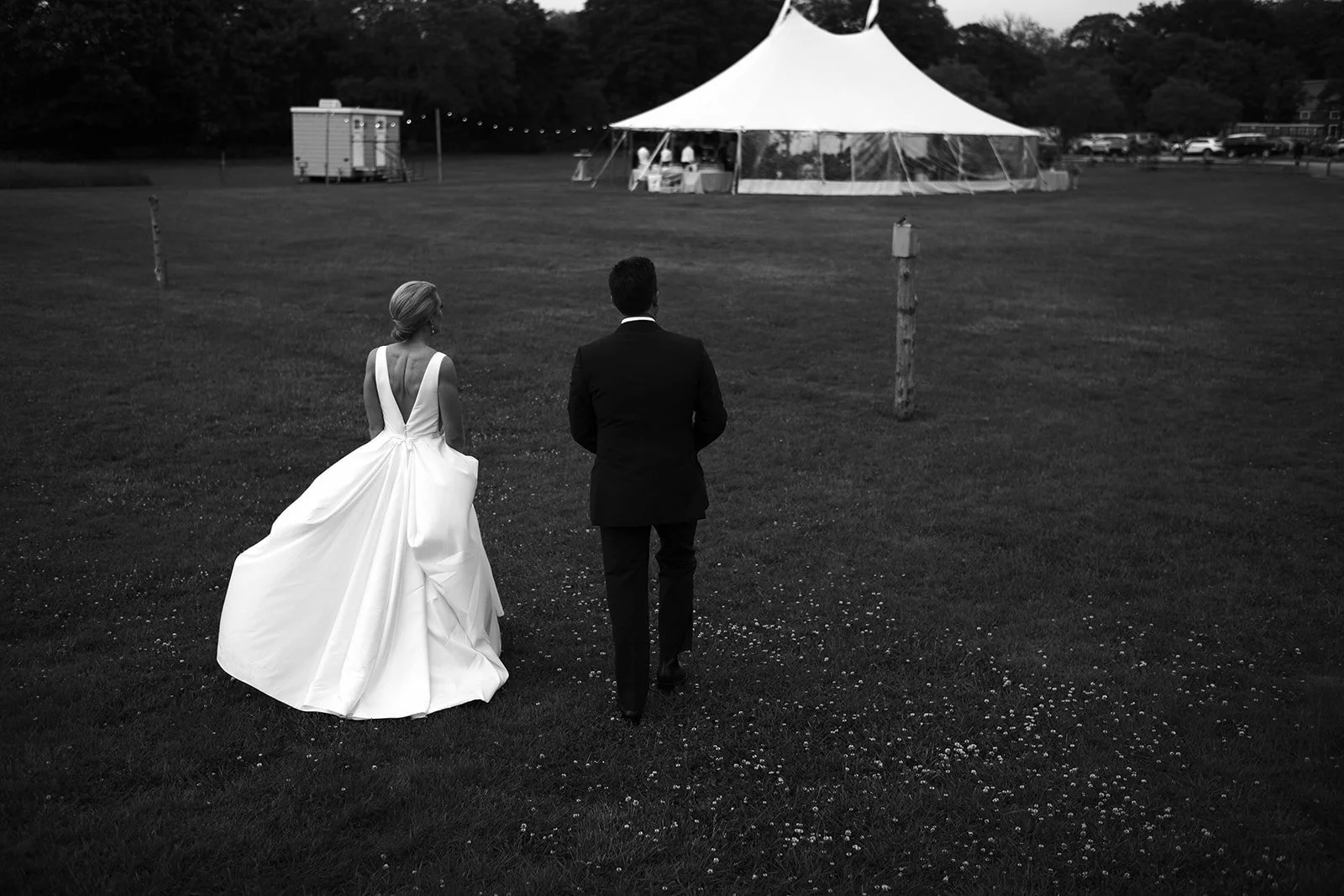 A bride in a wedding dress and a groom in a suit walking towards a wedding tent on a grassy field.