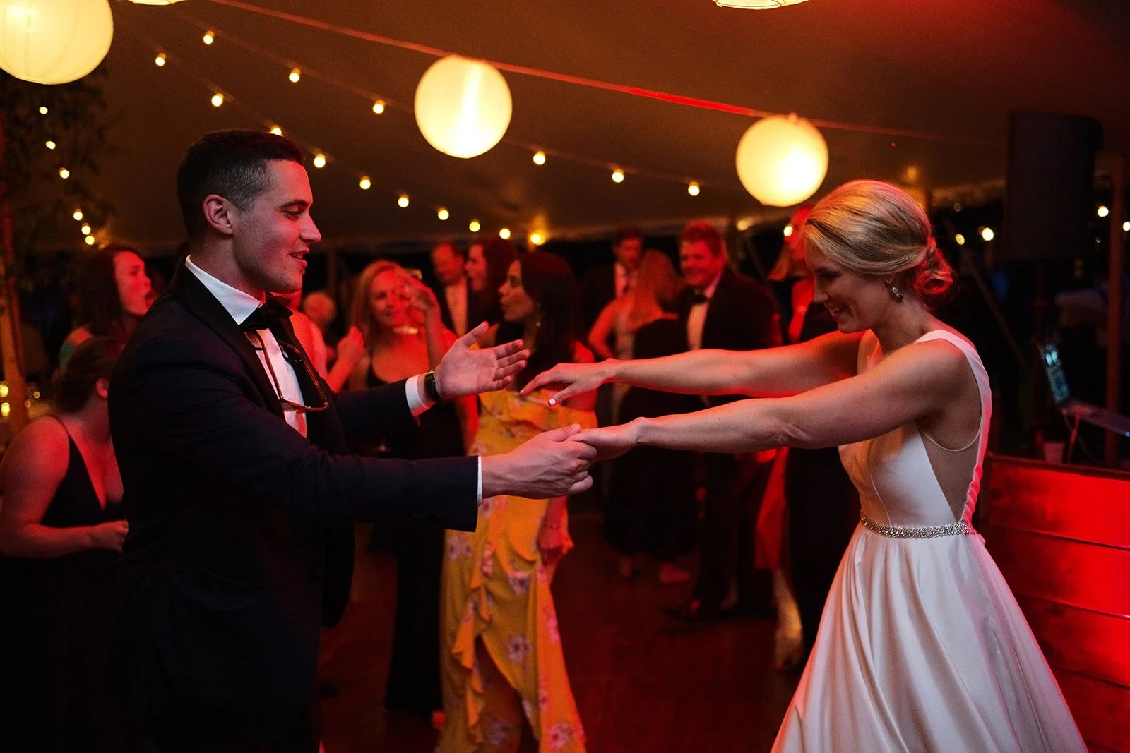 A bride and groom dancing at their wedding reception, surrounded by guests under hanging paper lanterns and string lights.