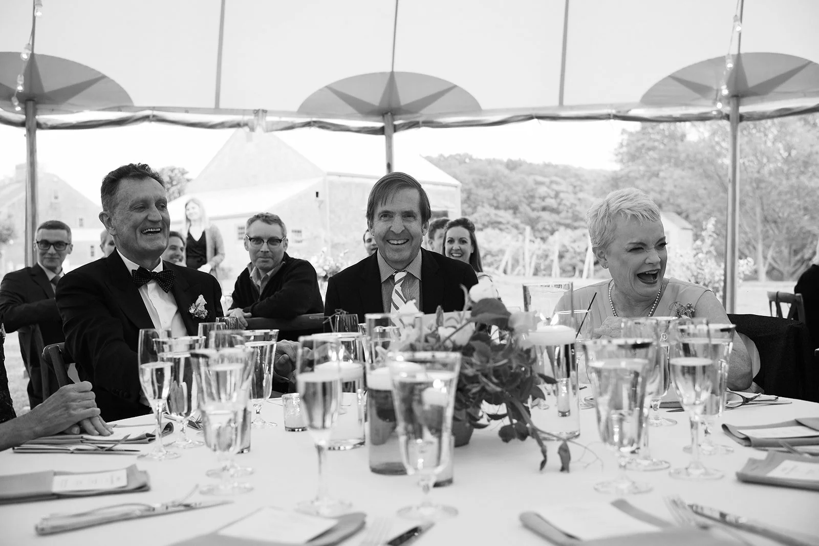 Black and white photo of a group of people sitting at a banquet table during a celebration, smiling and laughing, with glasses of water and table settings in the foreground and a tent and outdoor scenery in the background.