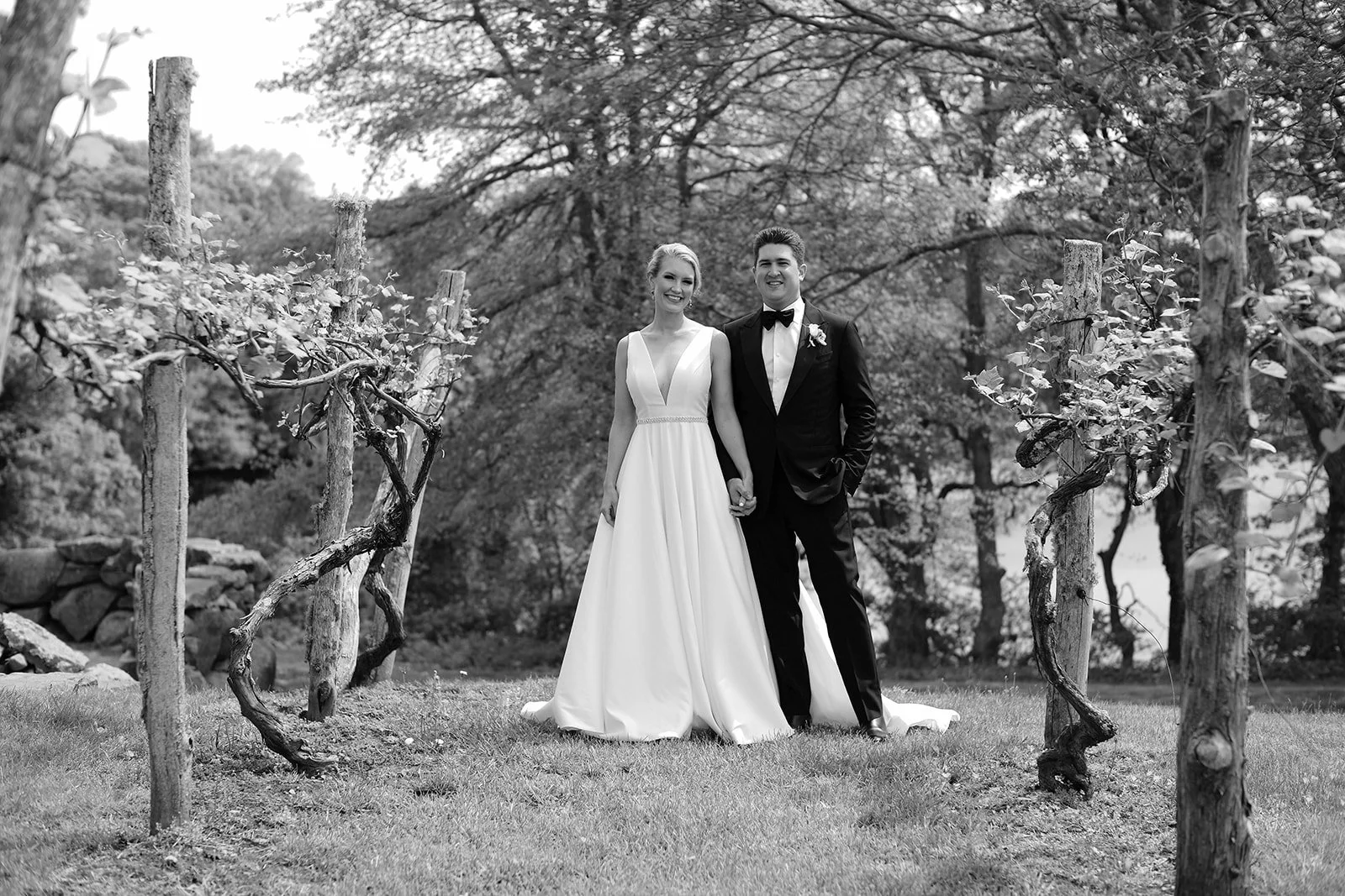 Black and white photograph of a bride and groom standing hand in hand outdoors among vineyard posts and vines, with trees in the background, smiling at the camera.