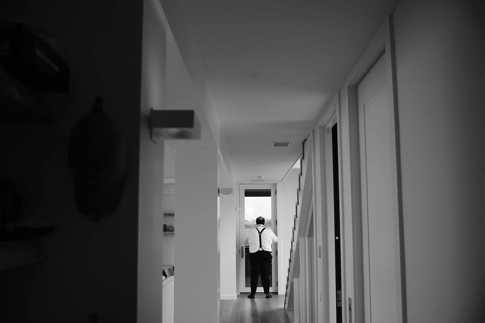A man wearing a white shirt and suspenders stands in a modern hallway, looking out a door at a landscape beyond.