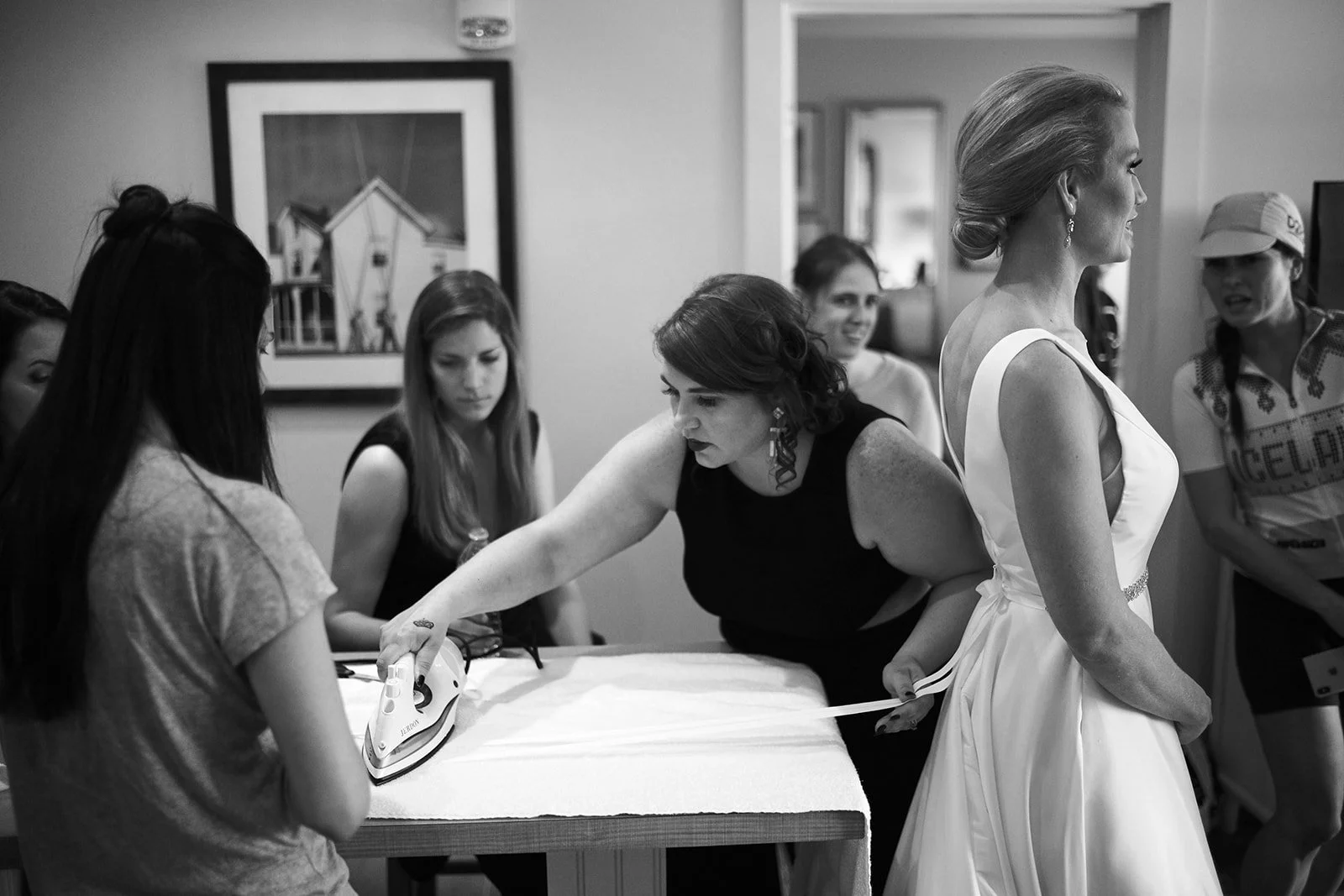 Women preparing for a wedding, with one woman ironing a dress on a table, amid other women gathered around in a room.