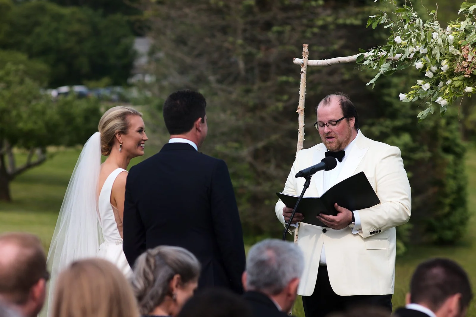 A couple getting married outside, facing an officiant who is reading from a book during the ceremony. The bride is wearing a white dress and veil, while the groom is in a dark suit. The officiant is dressed in a white tuxedo and glasses, holding a mi
