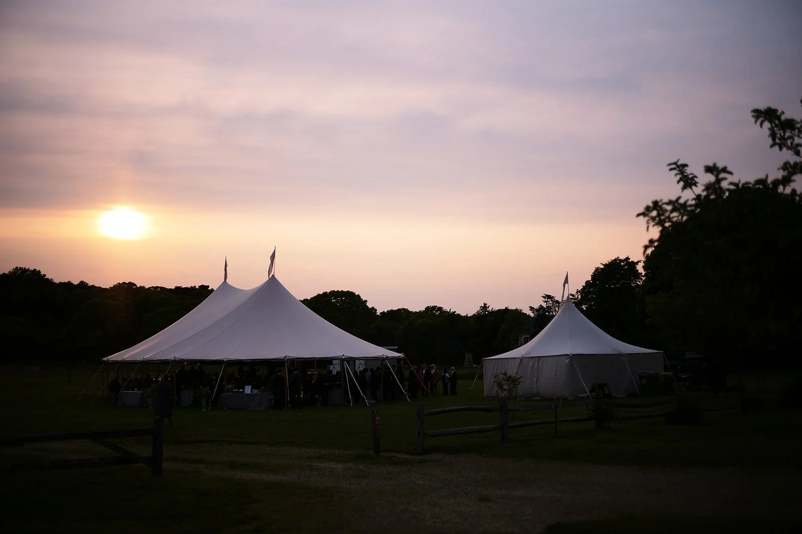 Two large white event tents set up outdoors during sunset with trees and cloudy sky in the background.