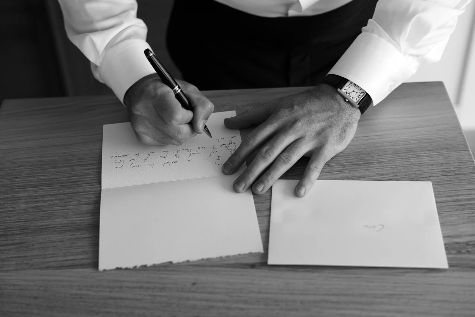 A person in a white shirt writes a letter with a pen on a piece of paper on a wooden table.