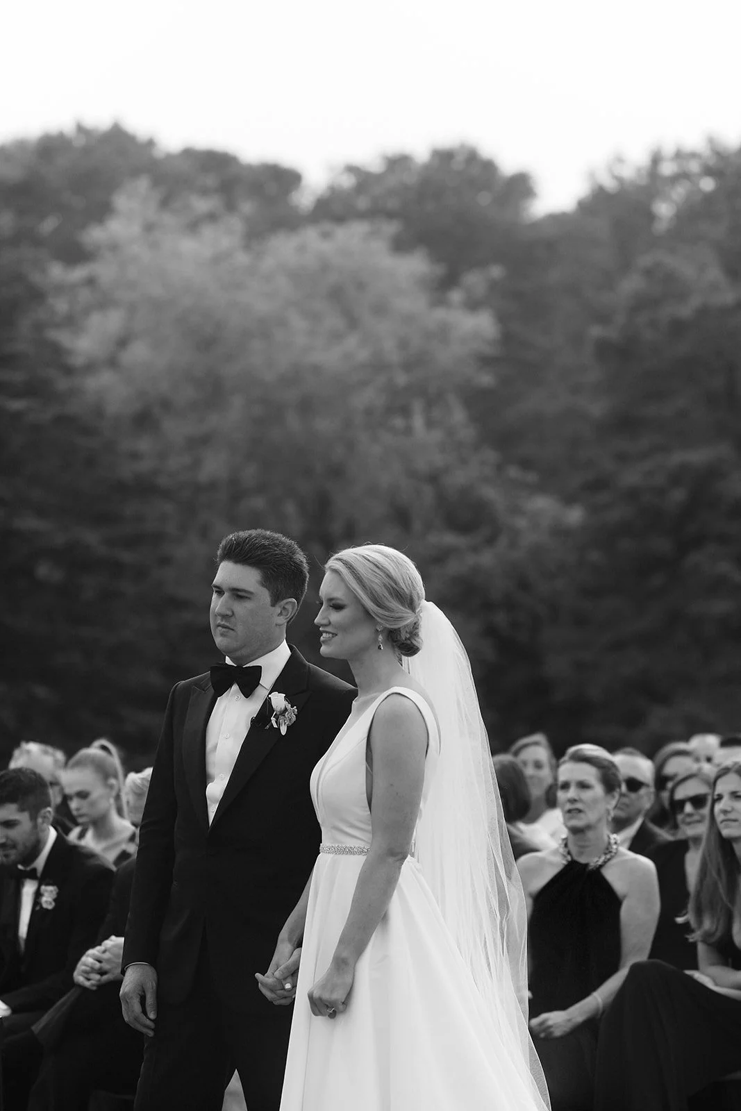 Black and white wedding photo of a bride and groom holding hands during ceremony, surrounded by seated guests outdoors.