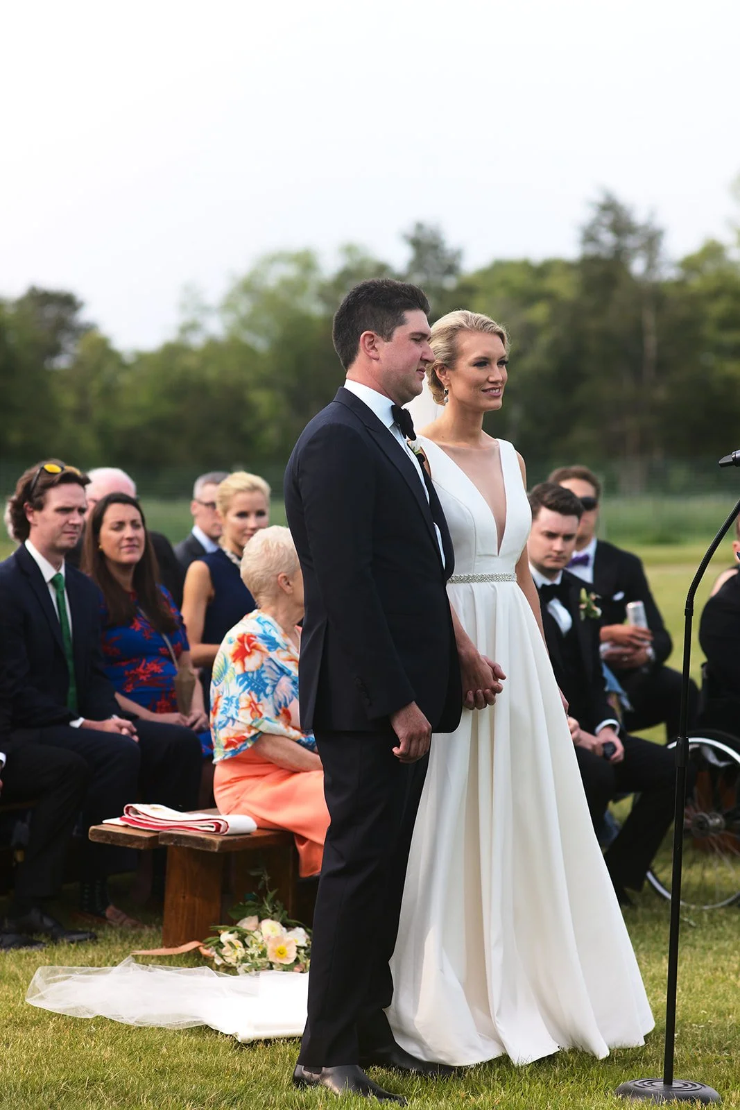 A couple is getting married outdoors, holding hands and standing in front of seated guests, with trees and a cloudy sky in the background.