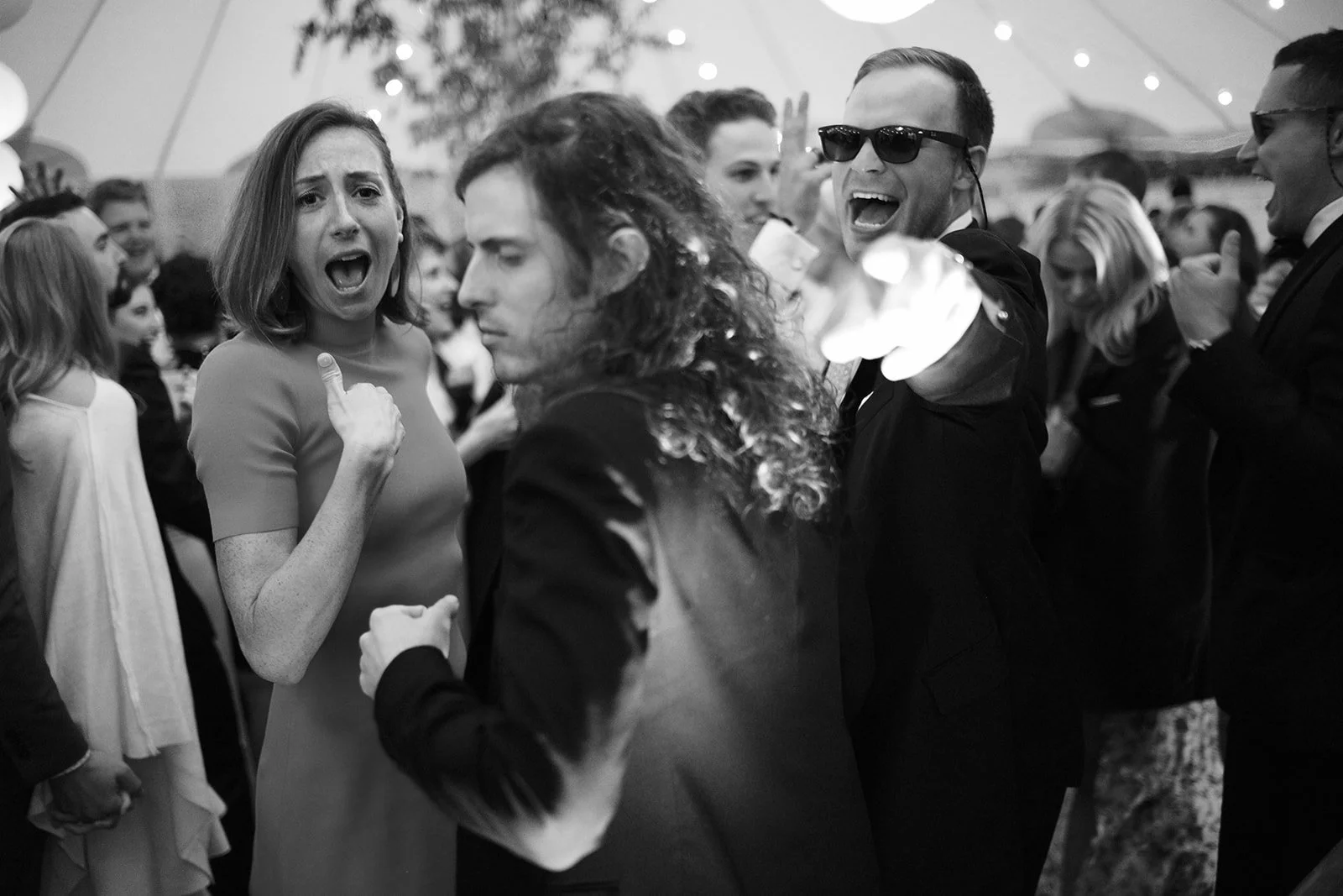 A group of people at a party or celebration, some dancing and others talking, with a woman in a dress and a man with sunglasses in the foreground, in a tent with string lights.