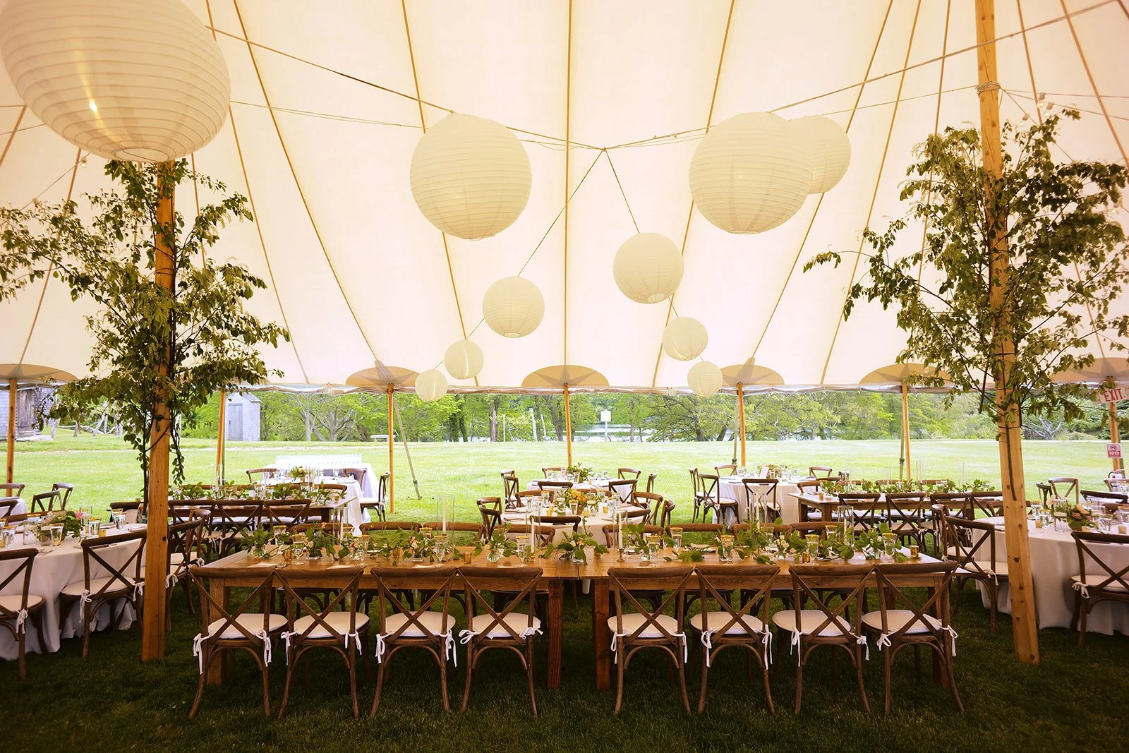 Interior of a large outdoor event tent decorated with paper lanterns, greenery, and tables set for a gathering on a grassy field.