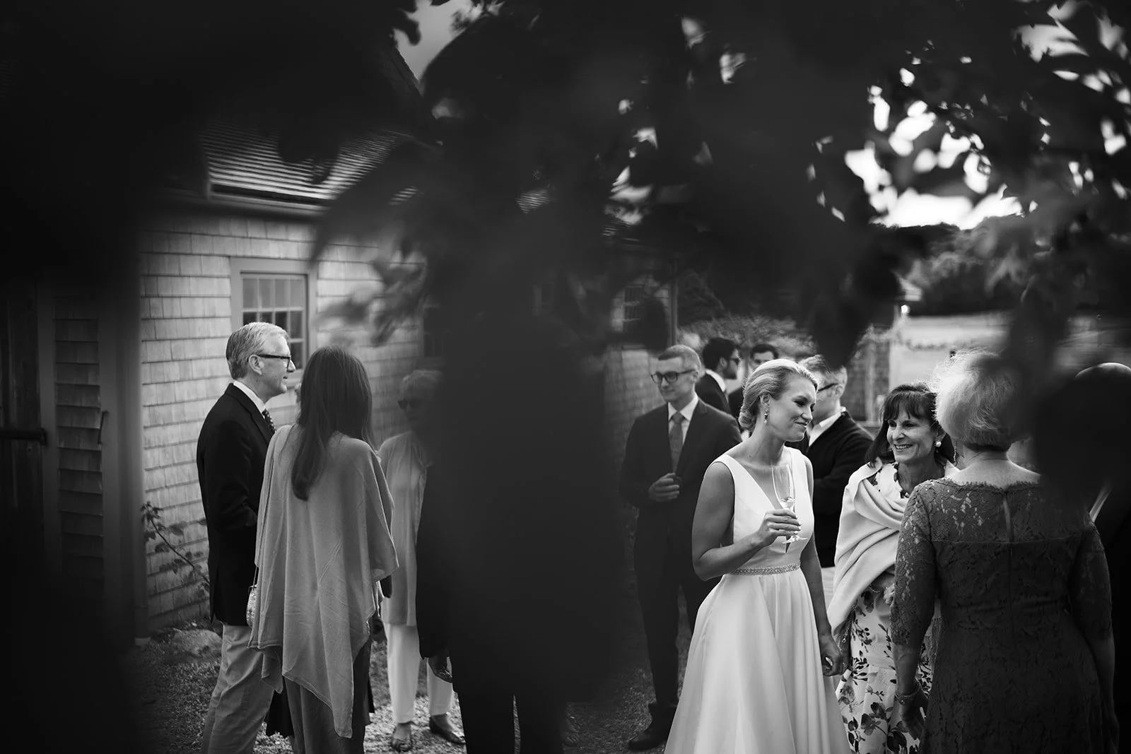 Black and white photo of a group of people at an outdoor gathering, with some holding glasses and engaging in conversation, a woman in a white dress holding a glass is prominent in the center.